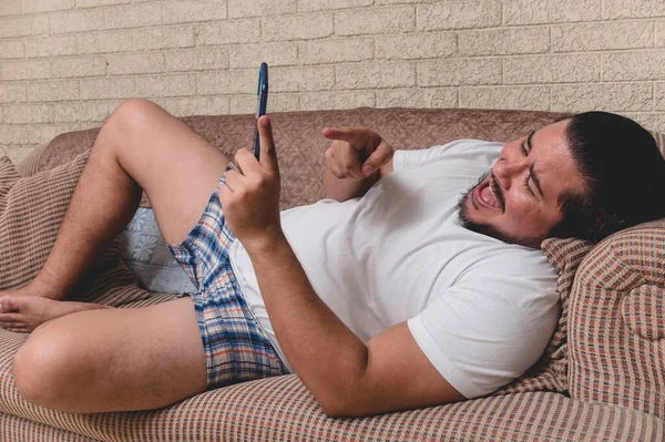 A overweight and unshaven man laughs at some funny videos streaming on the cellphone while lounging on the sofa during a lazy day.