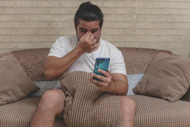 Karışık ırktan bir adam kanepede oturup telefonuna bakarken sinüs boşluğuna masaj yapıyor. Sinüzit ya da benzer bir sorun.