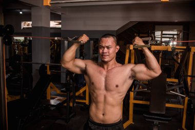 Formda ve kaslı Asyalı bir adam, sert bir spor salonunda iki kaslı kablo kıvırcık yapıyor. Kuvvetli bir şekilde pazı çalışıyor..