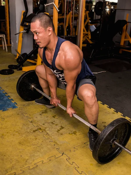 A large and muscular asian man does sumo barbell deadlifts at a hardcore gym. Working out quadriceps, glutes, hamstrings, and other muscles of the posterior chain.