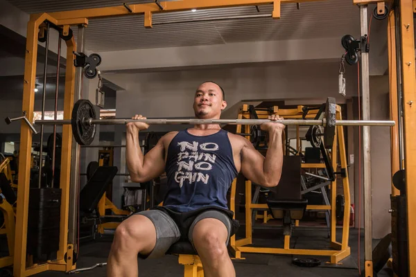 An athletic and rugged asian man does Seated Military Presses. Shoulder workout at the gym.