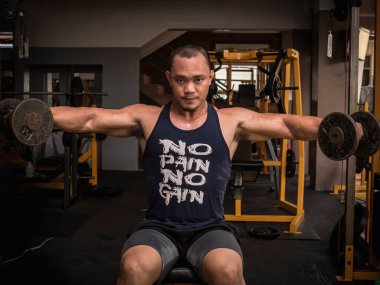 A large muscular asian man does seated dumbbell lateral raises with both arms. Shoulder workout at a hardcore gym. clipart