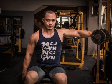 Büyük kaslı Asyalı bir adam bir kolunun dambılı iki koluyla kaldırır. Sıkı bir spor salonunda omuz egzersizi..