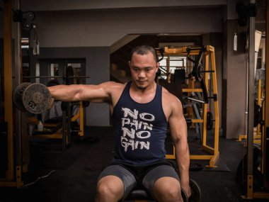 Büyük kaslı Asyalı bir adam iki koluyla bir kolunda dambıl kaldırıyor. Sıkı bir spor salonunda omuz egzersizi..