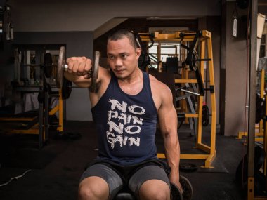 Büyük kaslı Asyalı bir adam tek koluyla bir dambıl kaldırıyor. Sıkı bir spor salonunda omuz egzersizi..