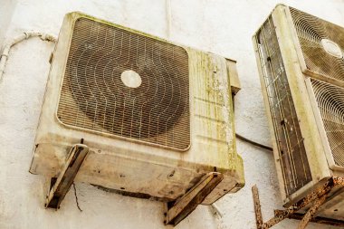 An old and aging Split type air conditioner condenser mounted on the exterior wall. Grimy and covered in slight moss and rust. Rusting brackets.