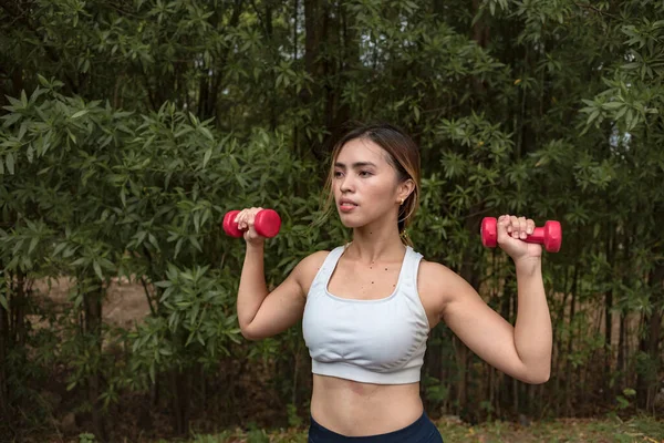 一位健康而纤细的亚洲女士站在一个自然公园里按着哑铃 肩部运动训练 — 图库照片