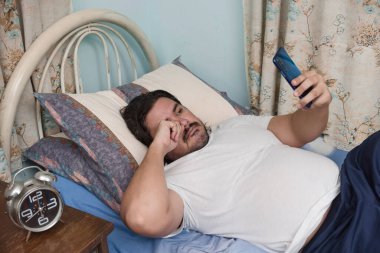 Bir adam cep telefonunda mesaj var mı diye kontrol ediyor. Yarı uykulu ve yatakta yatıyor. Yorgun gözlerini ovuşturarak uyandı..