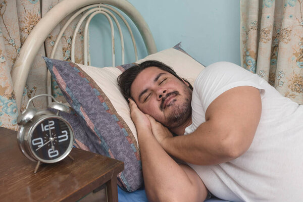 A bearded man of mixed race sleeping soundly on his side on the bed at his apartment. REM sleep.