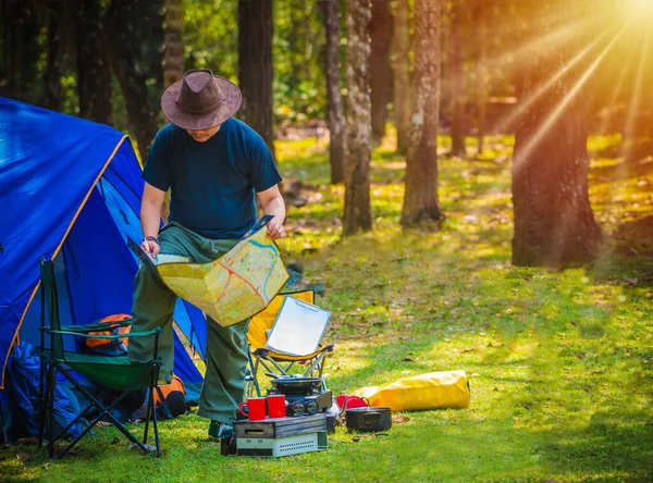 A man at the camping area. He is looking at a map with his notepad on a yellow chair. Bag packer with outdoor kitchen equipment and lantern, mug and tent. Copy space.