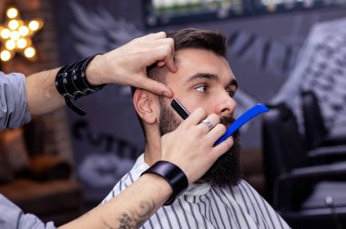 Genç sakallı adam modern berber dükkanında profesyonel olarak saçını kestiriyor. Sakal ve favoriler tıraş olurken ve klasik usturayla şekillendirilirken. Berber dükkanı reklam konsepti. Kasım Taşı.