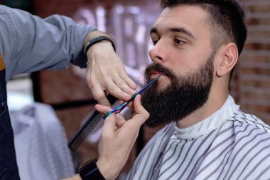 Sakal şekillendirme ve makasla kesme. Genç sakallı hippi berber dükkanında berber tarafından saç ve sakal kestiriliyor. Berber dükkanı reklam konsepti. Kasım Taşı.