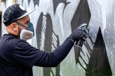 Graffiti sanatçısı iş başında, duvara sprey boyayla resim çiziyor, koruyucu maske takıyor ve filtreli solunum cihazı takıyor. Sokak sanatı kültürü kavramı.