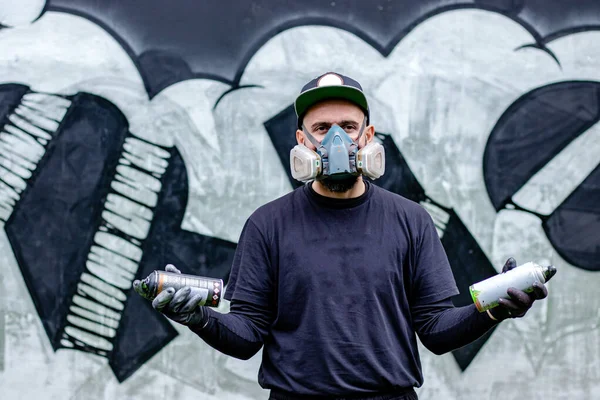 Graffiti sanatçısı, duvardaki çiziminin önünde koruyucu maske ve filtreli solunum cihazı takan iki sprey boyayla poz veriyor. Sokak sanatı kültürü kavramı.