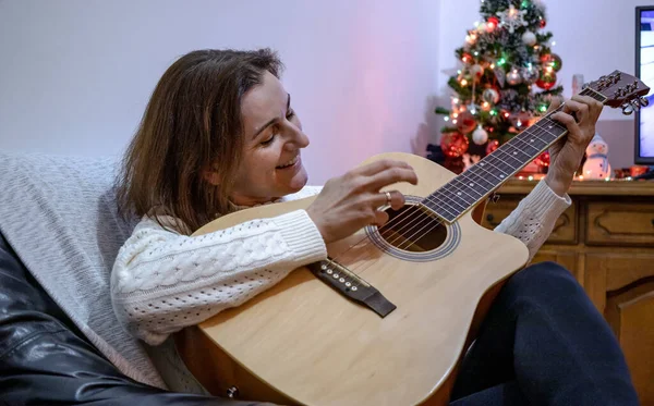 年轻女子在圣诞树前弹奏吉他 因季节假日被隔离而单独度过的时间 — 图库照片