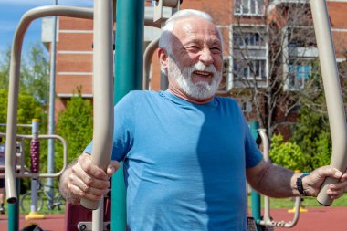 Yaşlı, gri sakallı bir adam açık havada spor aletleri üzerinde egzersiz yapıyor.. 