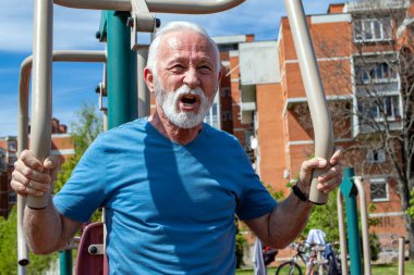 Yaşlı, gri sakallı bir adam açık havada spor aletleri üzerinde egzersiz yapıyor..