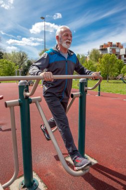 Yaşlı, gri sakallı bir adam açık havada spor aletleri üzerinde egzersiz yapıyor..