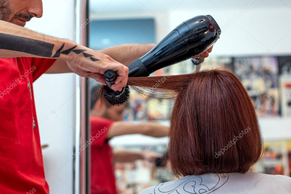 Brunette woman customer getting her hairstyle by a professional hairstylist at a beauty salon. Close up of a hairdresser's hands with a hairdryer and a brush. 