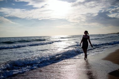 Mavi okyanus ya da deniz sahillerinde büyük dalgalarla yürüyen genç bir kadının silueti..