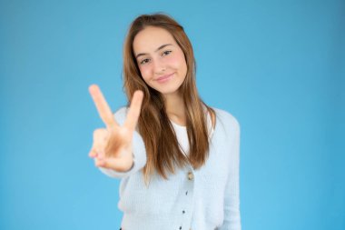 Gülümseyen mutlu bir kadının portresi zafer işareti gösteriyor ve mavi arka planda izole edilmiş kameraya bakıyor.