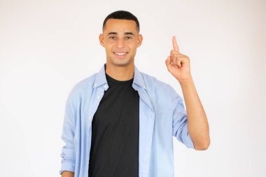 Mavi gömlekli çekici bir genç, beyaz arka planda, parmağını kaldırarak işaret ediyor.