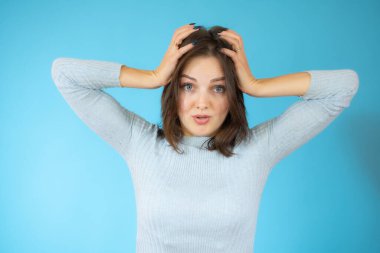 Genç, güzel bir kadın kendini stresli, endişeli, endişeli ya da korkmuş hissediyor, elleri başının üstünde, mavi arka planda hata yaparken panikliyor..