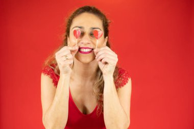 Aşkımı al! Çekici genç bir kadın kırmızı bir kalp tutuyor ve kırmızı arka planda tek başına dikilirken kameraya gülümsüyor.