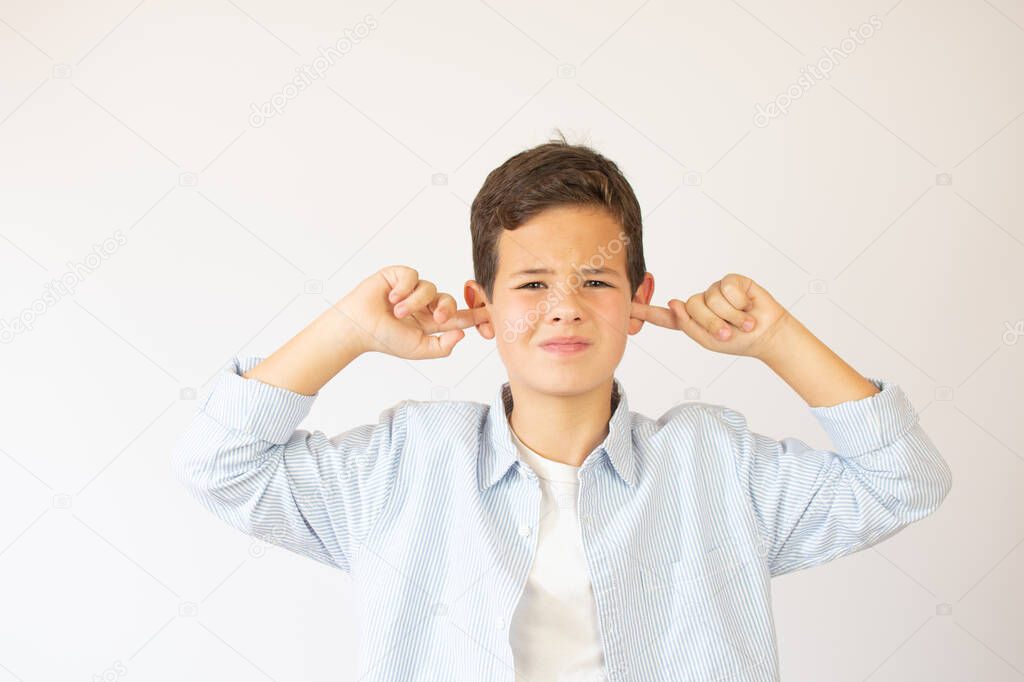 Don't want to hear! Angry young boy in stylish shirt with hood closing ears with fingers, disobedience, ignoring annoying noise. Indoor studio shot isolated on white background