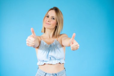 Mutlu, genç bir kadın mavi arka planda başparmak kaldırıyor..