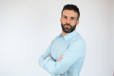 Kollarını kavuşturup beyaz arka plan üzerinden kameraya bakan sıradan mavi gömlekli genç bir adam..