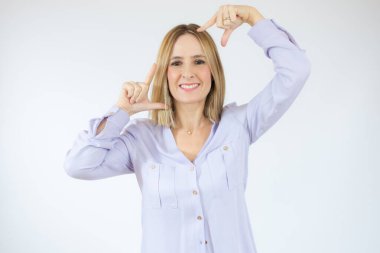 Genç ve güzel bir kadın izole edilmiş beyaz arka planda duruyor gülümsüyor ve elleri ve parmakları gülen yüzlü bir çerçeve yapıyor. Yaratıcılık ve fotoğrafçılık kavramı.