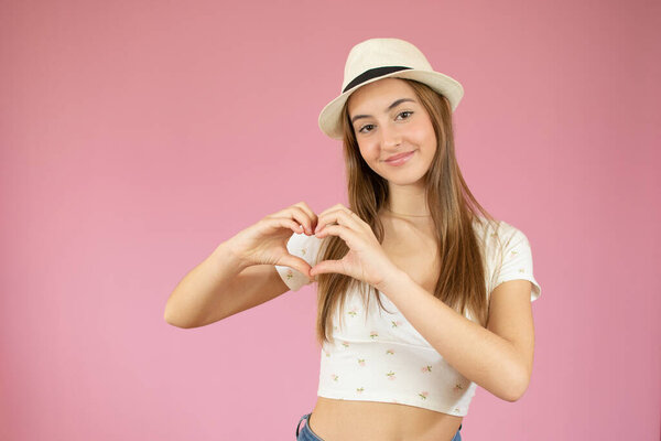 молодая девушка в футболке делает сердце символ руками на изолированном розовом фоне