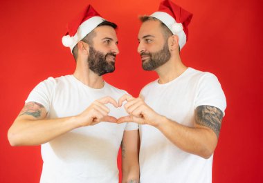 Mutlu eşcinsel çift Noel Baba şapkası ve günlük giysiler içinde, parmaklarından kalp şekli oluşturuyorlar, kırmızı arka planda stüdyo resmi yapıyorlar.