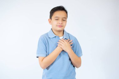 Güzel çocuk, mavi günlük tişört giyiyor, beyaz arka plan üzerinde duruyor, elleri göğsünde, gözleri kapalı ve yüzünde minnet dolu bir jest ile gülümsüyor. Sağlık kavramı.