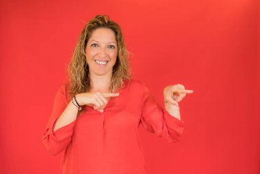 Gülümseyen kıvırcık kadın işaret ediyor ve kırmızı arkaplanda kameraya bakıyor.