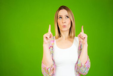 Genç bir kadın, yeşil arka planda dikilirken iki işaret parmağıyla işaret ediyor..