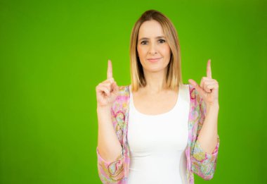 İki işaret parmağıyla işaret eden genç bir kadın kameraya neşeyle gülümserken yeşil arka planda duruyor..