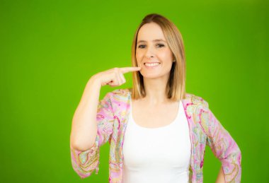 Yeşil izole edilmiş arka planda dikilen sıradan bir gömlek giyen genç ve güzel bir kadın. Gülümseyen, parmaklarıyla ve ağzıyla işaret eden. Diş sağlığı konsepti.