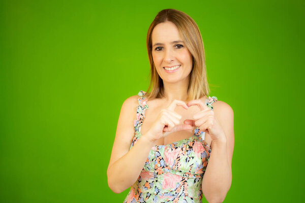 Красивая женщина показывает фигуру сердца с пальцами и улыбается в камеру на зеленом фоне