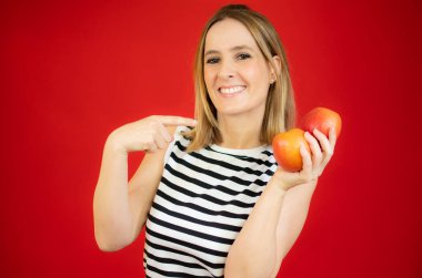 Kırmızı arka planda iki elma tutan çizgili tişörtlü, gülümseyen kadın..