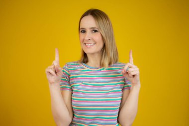 İki işaret parmağıyla işaret eden genç bir kadın kameraya neşeyle gülümserken, sarı arka planda duruyor..