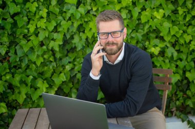 Diz üstü bilgisayarda çalışan ve dışarıda tahta masada otururken cep telefonuyla konuşan mutlu genç adam.