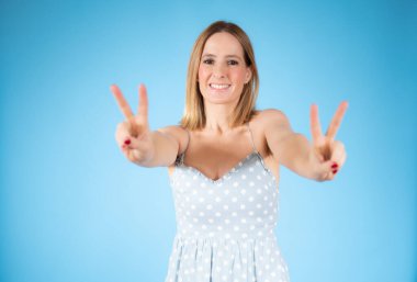 Genç ve güzel bir kadın kameraya bakıp mavi arka planda zafer işareti yapıyor. İki numara..