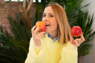 Sarı kazak giyen genç bir kadın, elinde iki elma tutuyor.