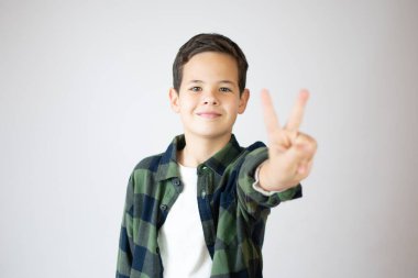 Beyaz duvarın önünde duran beyaz çocuk iki numaralı parmaklarıyla gülümseyerek kendinden emin ve mutlu görünüyor..