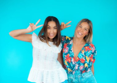 Stüdyoda, mavi arka plandaki kameraya bakarken zaferin sembolünü yapan iki kadının resmi.