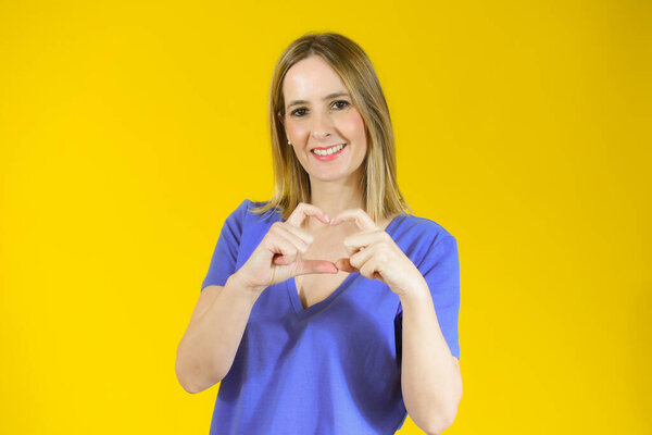 молодая красивая женщина показывает пальцем сердечную фигуру, посылая любовь своему любовнику, стоя на желтом фоне