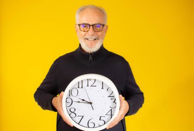 Sarı arka planda büyük bir duvar saati tutan yaşlı bir adamın stüdyo portresi.