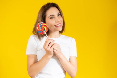 Elinde renkli lolipop şekeri tutan güzel genç bir kadın sarı arka planda eğleniyor. Tatlı yemek ve mutluluk konsepti.